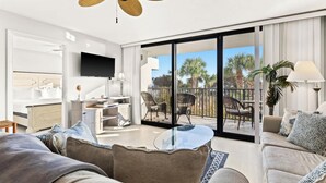 Ein modernes Wohnzimmer mit einer Couch, einem Couchtisch und einem Fernseher. Es gibt einen Balkon mit Stühlen und einer Brüstung.