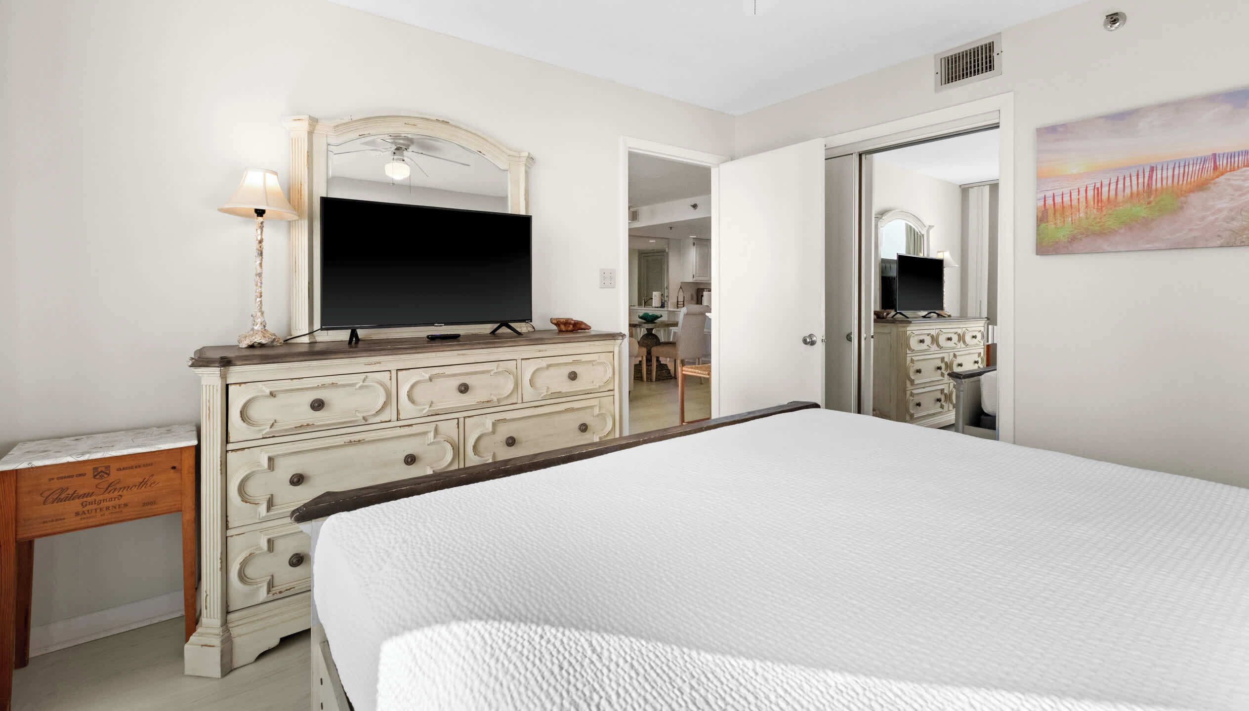 A bedroom with a large bed, a dresser with a mirror, a television, and a painting on the wall.