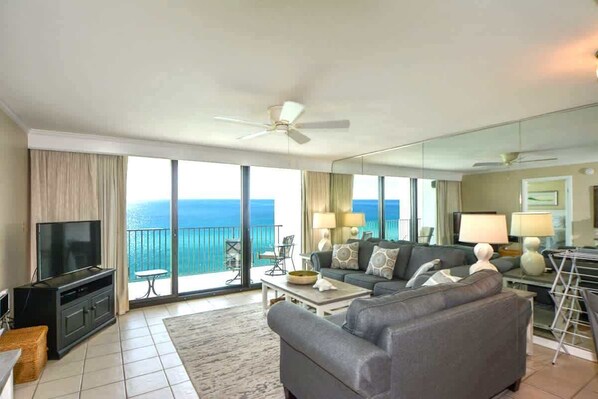 A spacious living room with a large flat-screen TV, a grey sofa, a coffee table, and a balcony with a view of the ocean. - One Seagrove Place 1205 (Santa Rosa Beach)
