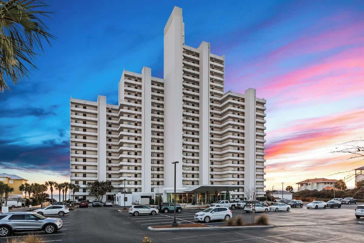 A multi-story hotel with a central tower, a parking lot with several cars, and palm trees in the foreground.