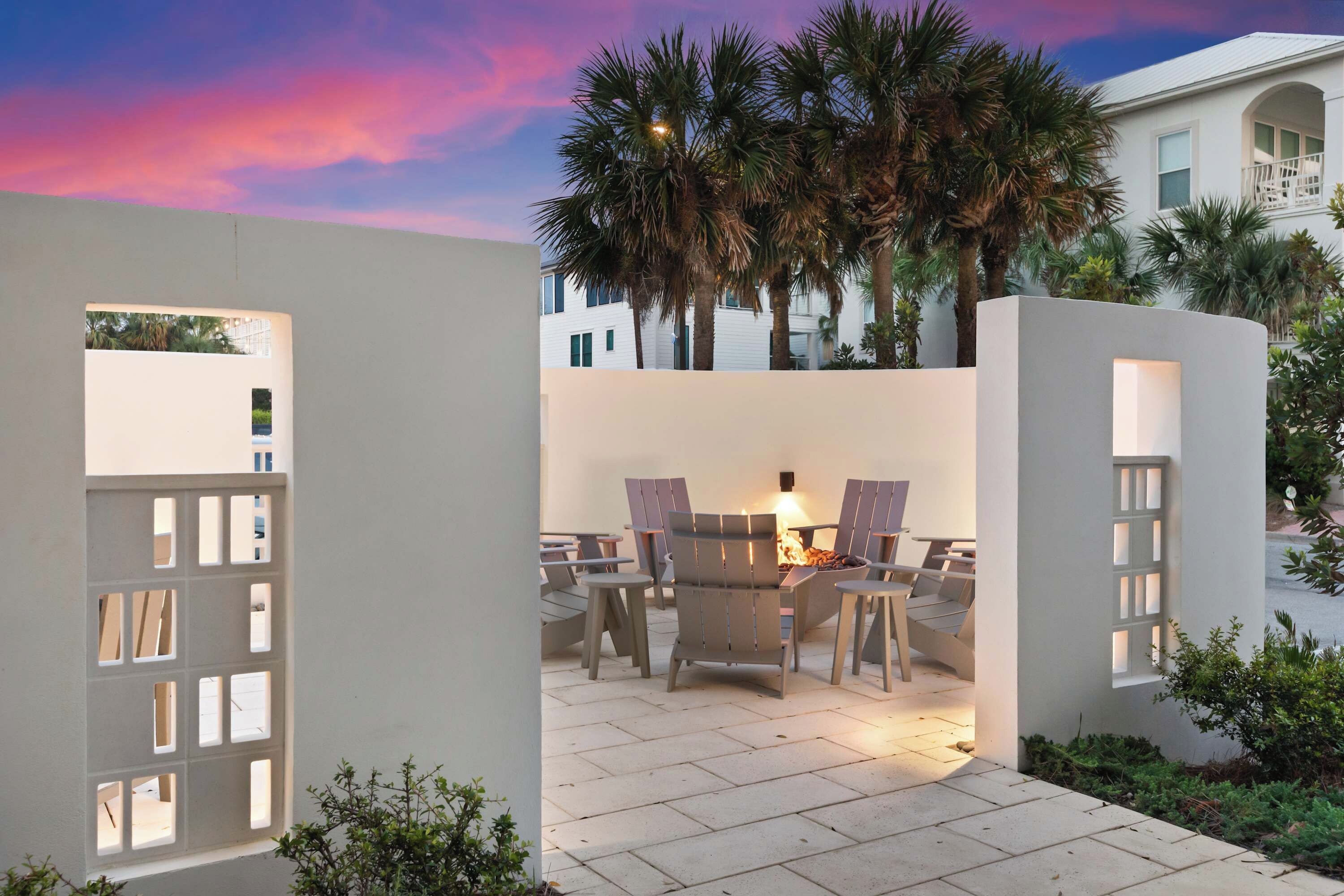 An outdoor patio area with a fire feature, seating, and a view of palm trees and a building in the background.