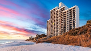 A high-rise hotel with a modern design, located adjacent to a beach with visible footprints.