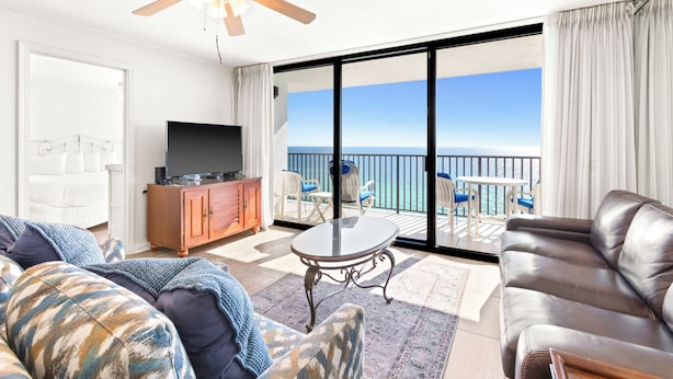 A living room with a TV, a sofa, a leather couch, and a balcony with a view of the ocean.