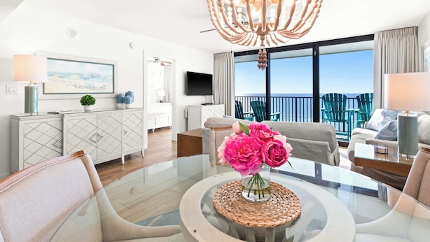 A modern living room with a glass coffee table, a sofa, and a TV. There is a balcony with a view of the sea.