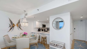 A modern kitchen with white cabinets, a microwave, and a coffee maker. There is a dining area with a table, chairs, and a chandelier. A round mirror hangs on the wall, and a door leads to another room.