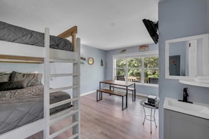 Une pièce avec des lits superposés, une petite table, un banc, une fenêtre donnant sur la verdure et un lavabo avec un miroir.