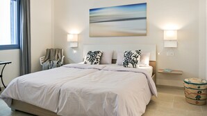 A bedroom with a large bed, a painting above the bed, two bedside lamps, a small table, a chair, and a decorative basket. - Holiday flat with balcony and sea view (La Caletta)
