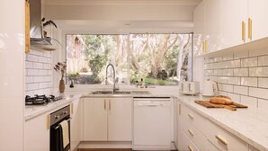 Een moderne keuken met witte kastjes, een inbouwvaatwasser, een gasfornuis en een groot raam met uitzicht op bomen en een tuin.