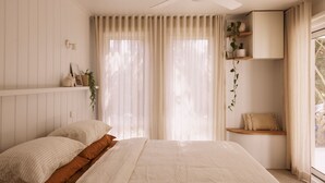 Een slaapkamer met een bed, een raam met dunne gordijnen, een plank met decoratieve objecten en uitzicht op de omgeving.