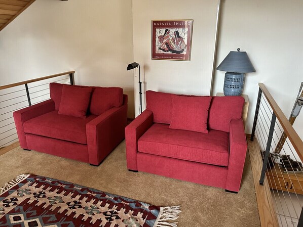 A living room with two red sofas, a patterned rug, a lamp, and a framed artwork on the wall.