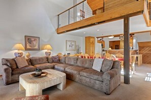 A spacious living room with a large sectional sofa, a coffee table, and a dining area with wooden chairs. A wooden staircase leads to an upper level.
