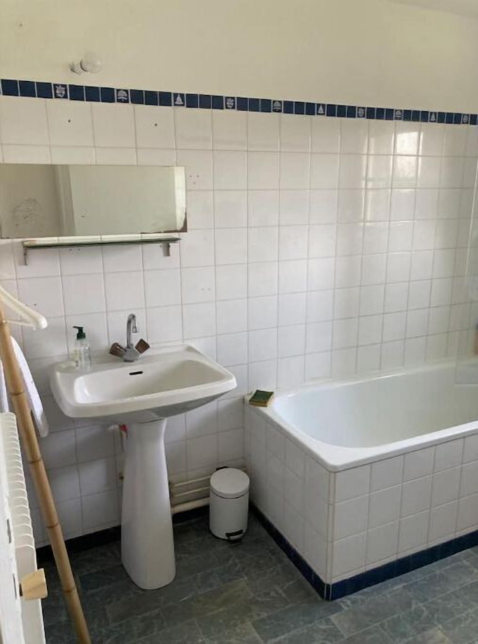 A bathroom with a pedestal sink, a mirror, a bathtub, and a trash can.