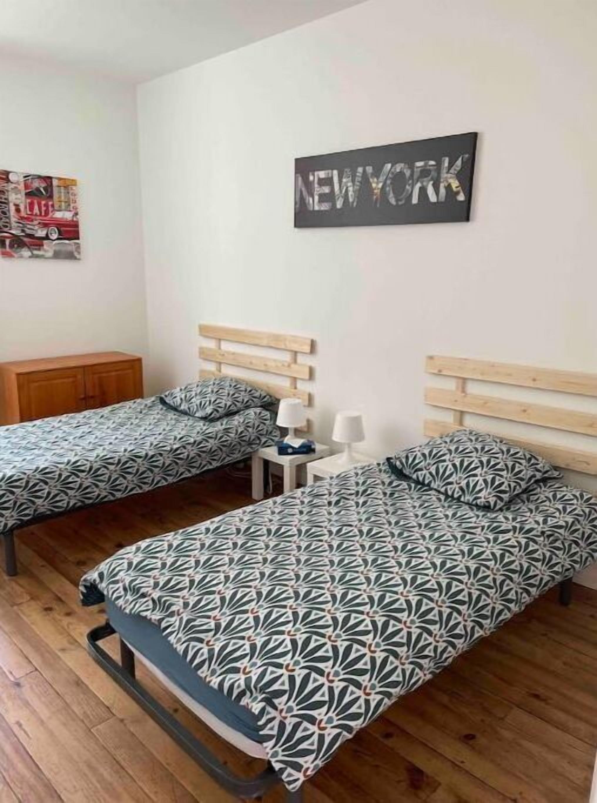 Ein Zimmer mit zwei Betten, beide mit gemusterten Bettdecken und passenden Kissen. Zwischen den Betten steht ein kleiner Tisch mit einer Lampe. An der Wand über den Betten hängt ein Plakat mit der Aufschrift "NEW YORK".