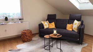 A cozy loft bedroom with a dark grey sofa, a small wooden coffee table, and a large window with a view of the city. - Moderne Ferienwohnung mit Atemberaubender Aussicht bei Heidelberg (Dossenheim)