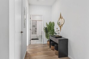 A hallway with a wooden floor, a black console table with decorative items, a potted plant, and a bathroom visible through an open door.