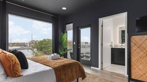 A modern hotel room with a large bed, a view of the city, a balcony, and a small bathroom.