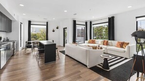 A modern living room with a white sofa, a dining area with a black table, and a kitchen with stainless steel appliances.
