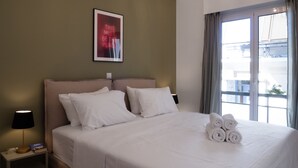 A hotel room with a large bed, a nightstand with a lamp, a framed picture on the wall, and a window with curtains. - Modern 1BR Apt, Next to Goulandris (Athens)