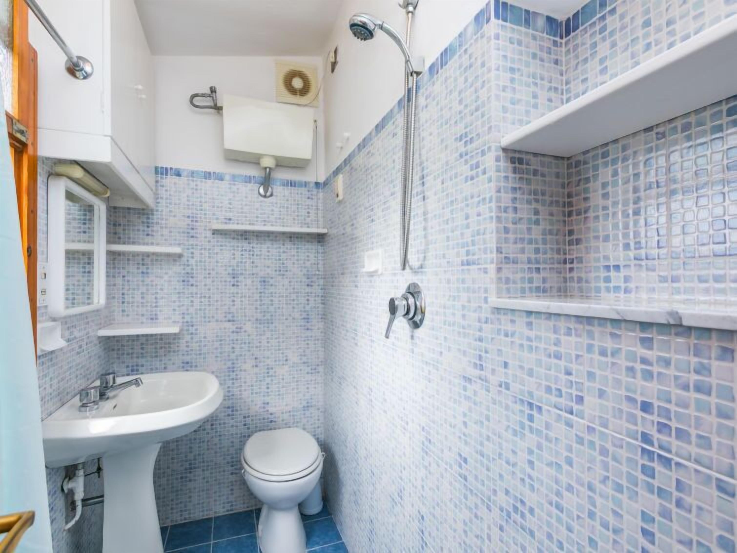 A bathroom with a white sink, toilet, and shower. The walls are tiled with blue mosaic tiles.