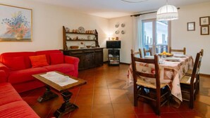 Un salon avec un canapé rouge, une table à manger en bois avec des chaises, un coin cuisine avec des étagères et une grande fenêtre avec des rideaux.