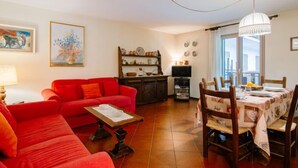 Un salon avec un canapé rouge, une table à manger avec des chaises, un téléviseur et un coin cuisine avec des étagères.