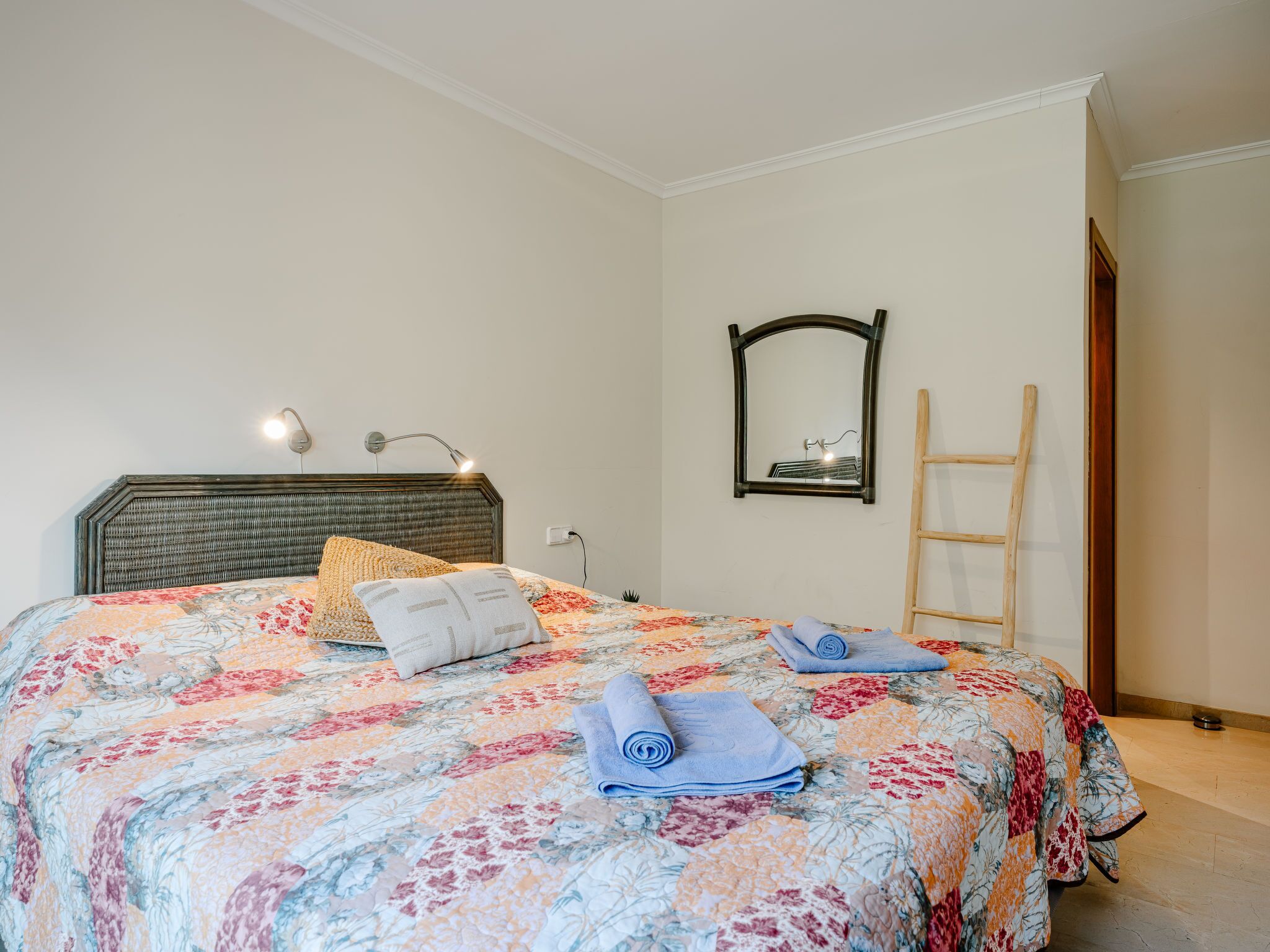 A neatly made bed with a floral-patterned bedspread, a headboard, and two rolled towels. There is a wall-mounted mirror and a wooden ladder leaning against the wall.