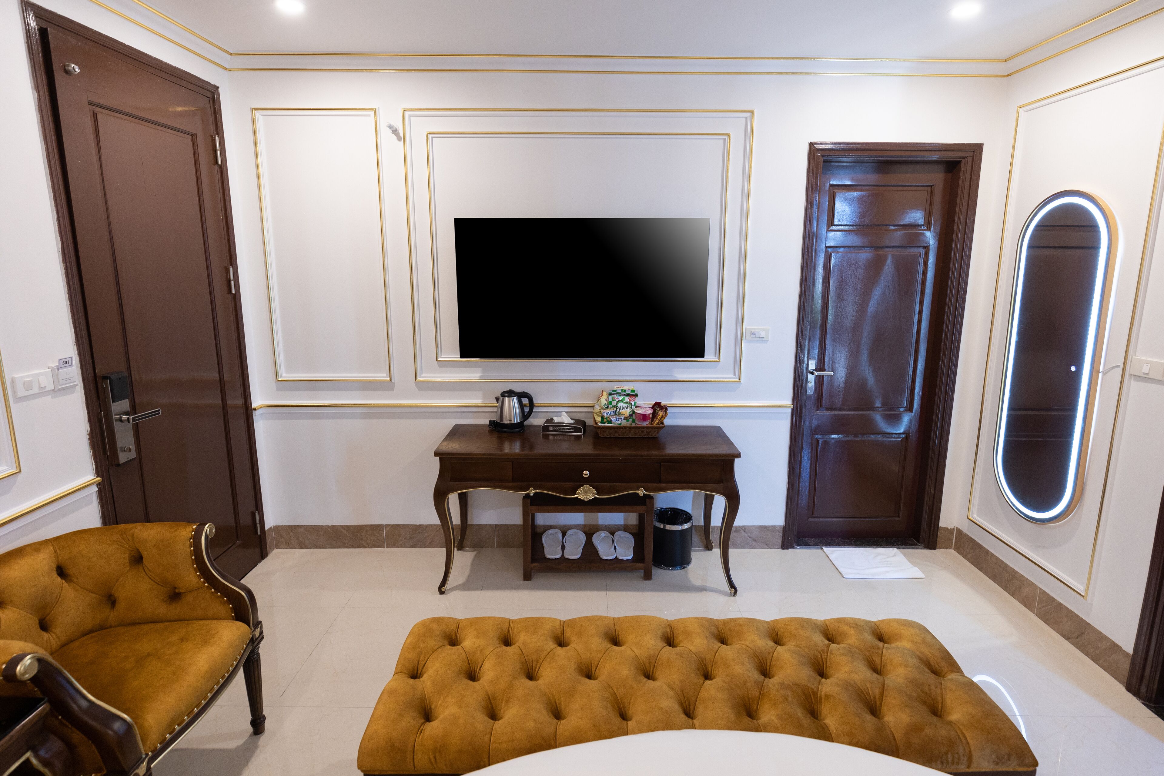 A room with a large flat-screen TV mounted on the wall, a wooden desk with a coffee pot, a pair of slippers, and a brown sofa.