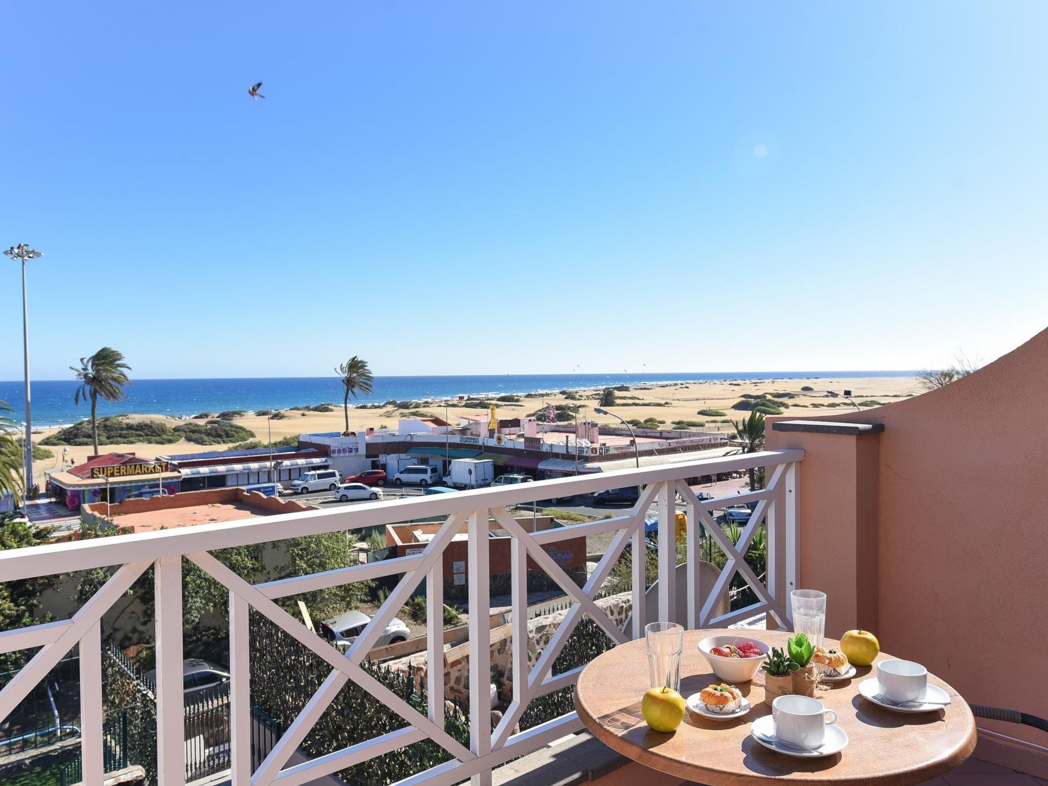 Un balcone con tavolo apparecchiato per la colazione, vista su una spiaggia e un supermercato.