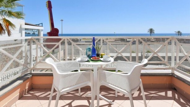 Un balcón con mobiliario de mimbre blanco, una guitarra, y una mesa dispuesta con una bebida y fruta.