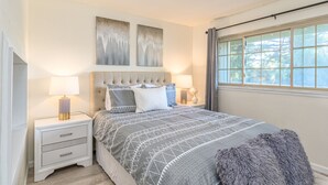 Een slaapkamer met een bed, twee nachtkastjes, een raam met gordijnen en twee aan de muur bevestigde kunstwerken.