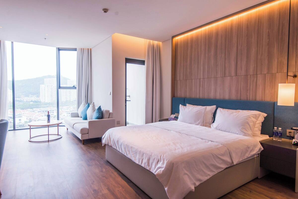 A modern hotel room with a large bed, a sitting area with a sofa and armchairs, a small table, and a view of the city through large windows.