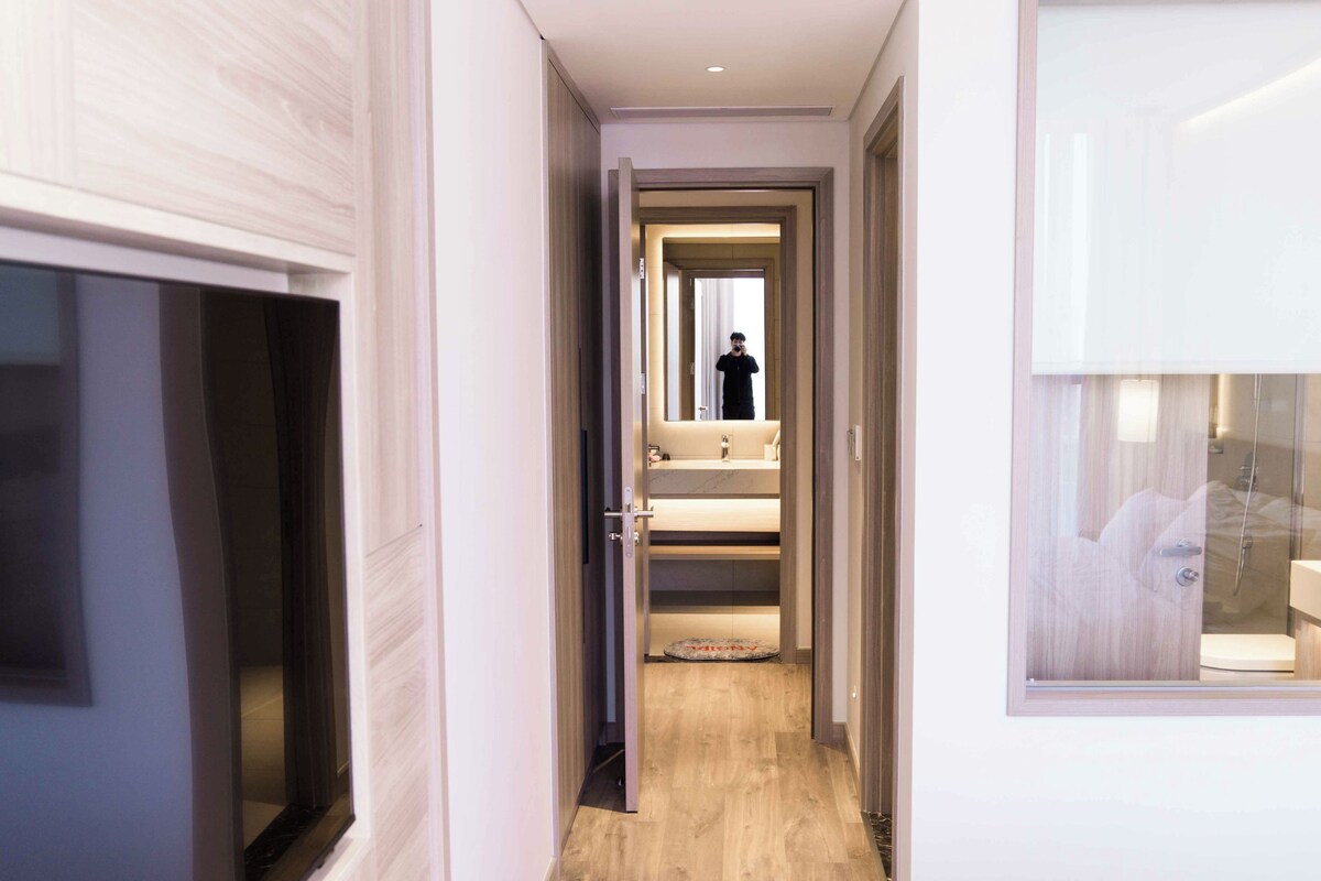 En moderne hotelgang med trægulv, et synligt badeværelse med bruser og et stort spejl, der reflekterer interiøret.