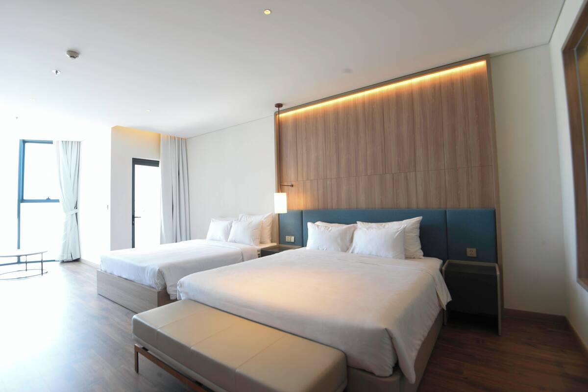 A modern hotel room with two beds, a bench, a wooden headboard, and a large window with curtains.