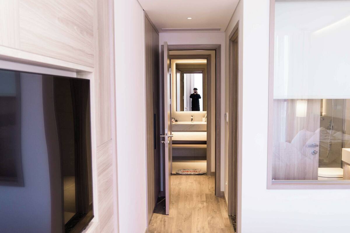 A modern hotel corridor with a wooden floor, a visible bathroom with a shower, and a large mirror reflecting the interior.