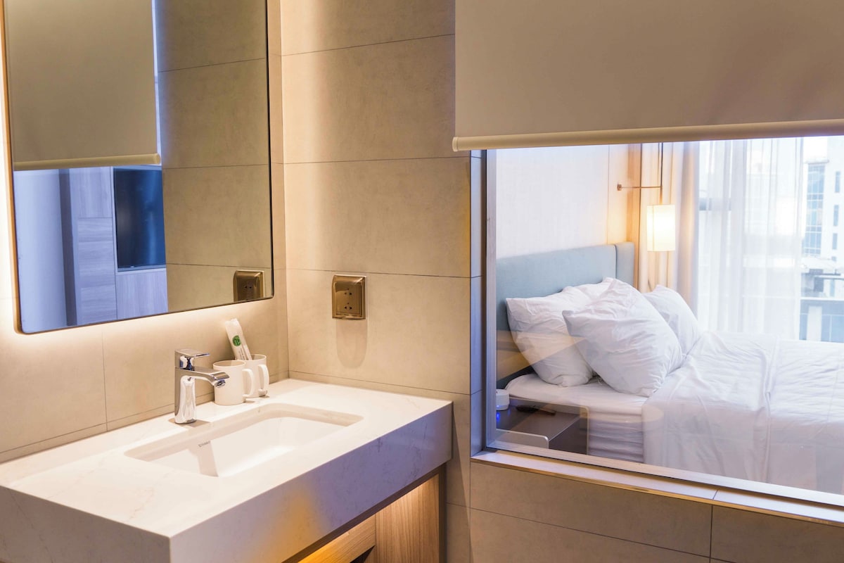 A modern hotel room with a large window, a bed with white linens, a bathroom with a sink and mirror, and a wall-mounted electrical outlet.