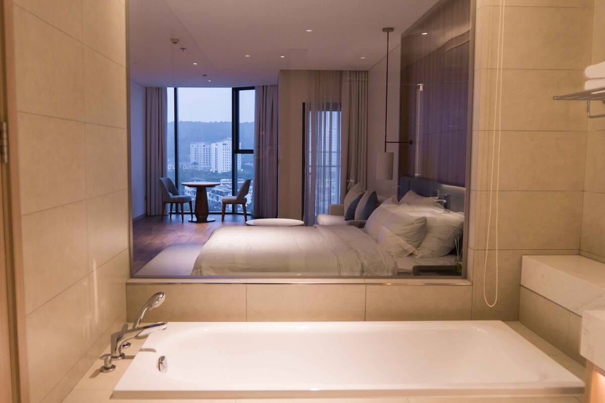 A modern hotel room with a large bed, a bathtub, and a view of a cityscape through large windows.