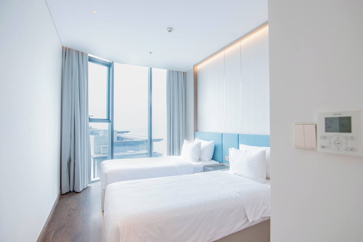 A modern hotel room with two beds, a large window with curtains, a view of a cityscape, and a control panel on the wall.