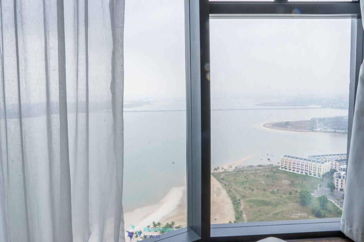 A view from a window with white curtains, overlooking a coastal area with buildings and a beach.