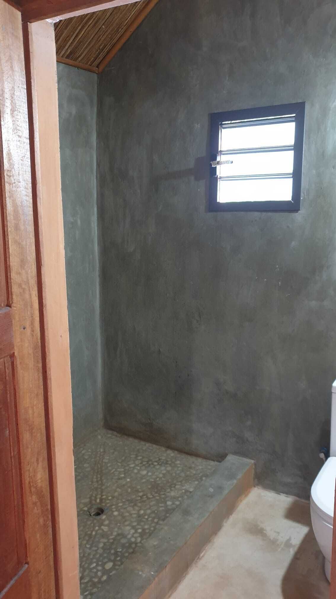 Een badkamer met een betonnen muur, een klein raam, een toilet en een douche.