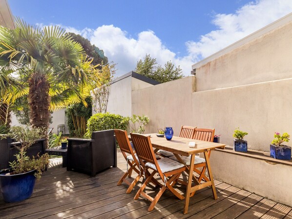 An outdoor patio with a wooden table and chairs, surrounded by potted plants and a palm tree.