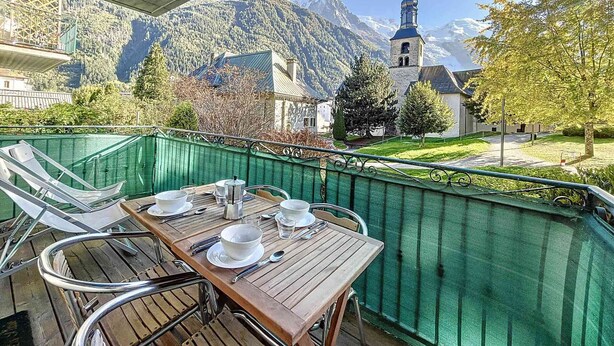 A balcony with a table set for two, overlooking a mountain landscape with a church. - Apartment in central Chamonix - 6 people, parking, 3-star comfort (Chamonix-Mont-Blanc)
