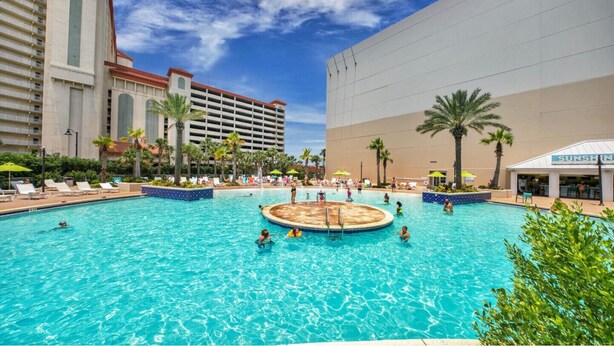 A large outdoor swimming pool with multiple lanes, surrounded by lounge chairs and palm trees.