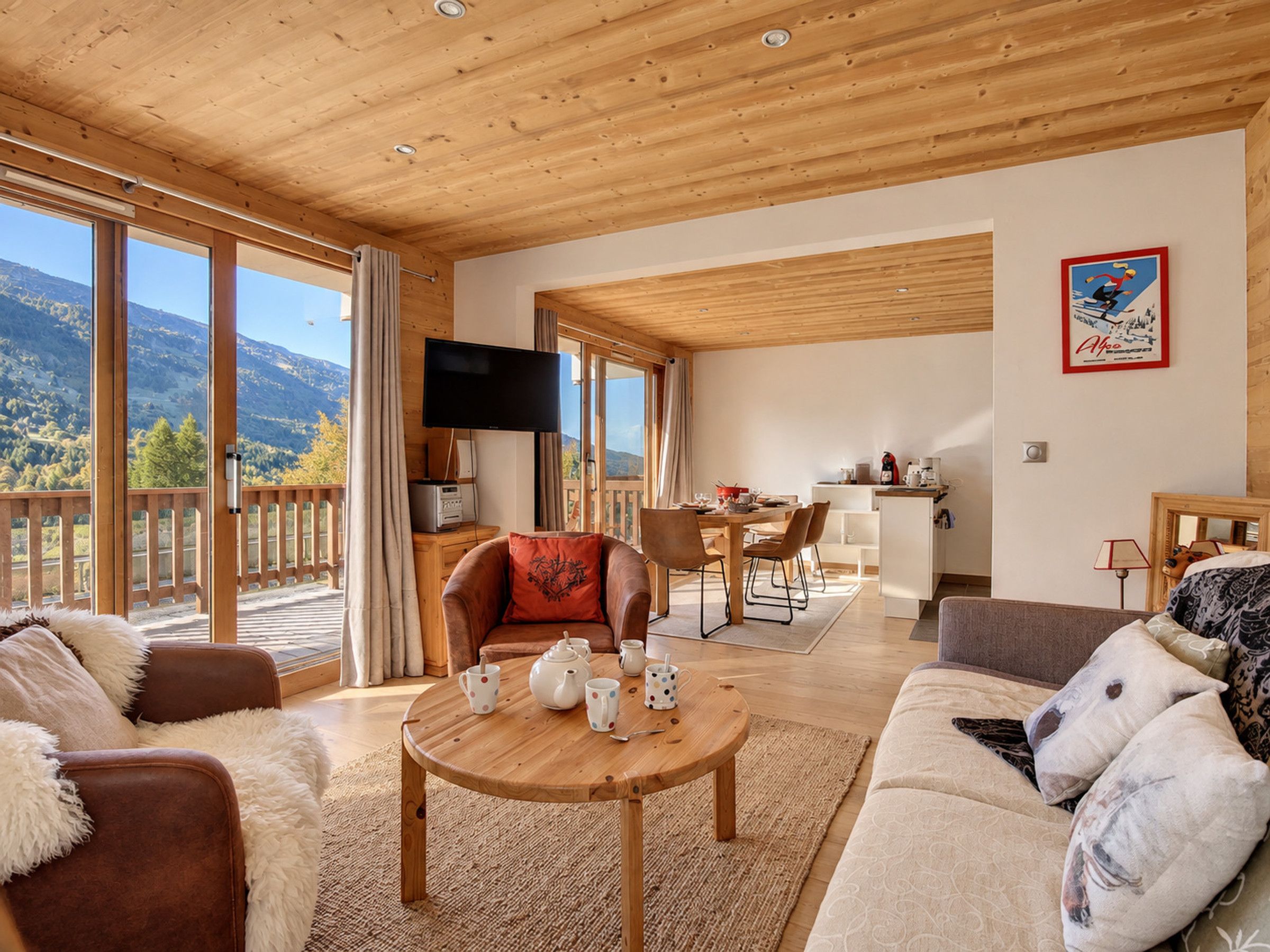 A spacious living area with a wooden ceiling, a large window offering a mountain view, a television mounted on the wall, and a dining area with a table and chairs.