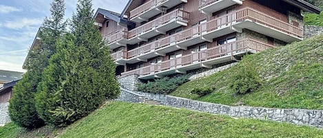 A multi-story hotel with balconies, set on a hillside with a stone retaining wall and greenery.