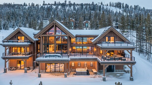 A large, multi-story wooden cabin with a central glass facade, surrounded by snow-covered trees. - Newly Built | Ski-in/Ski-out | Hot Tub (Big Sky)