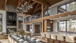 A spacious dining area with a long table set for a meal, a kitchen with modern appliances, and a large stone fireplace. - Newly Built | Ski-in/Ski-out | Hot Tub (Big Sky)