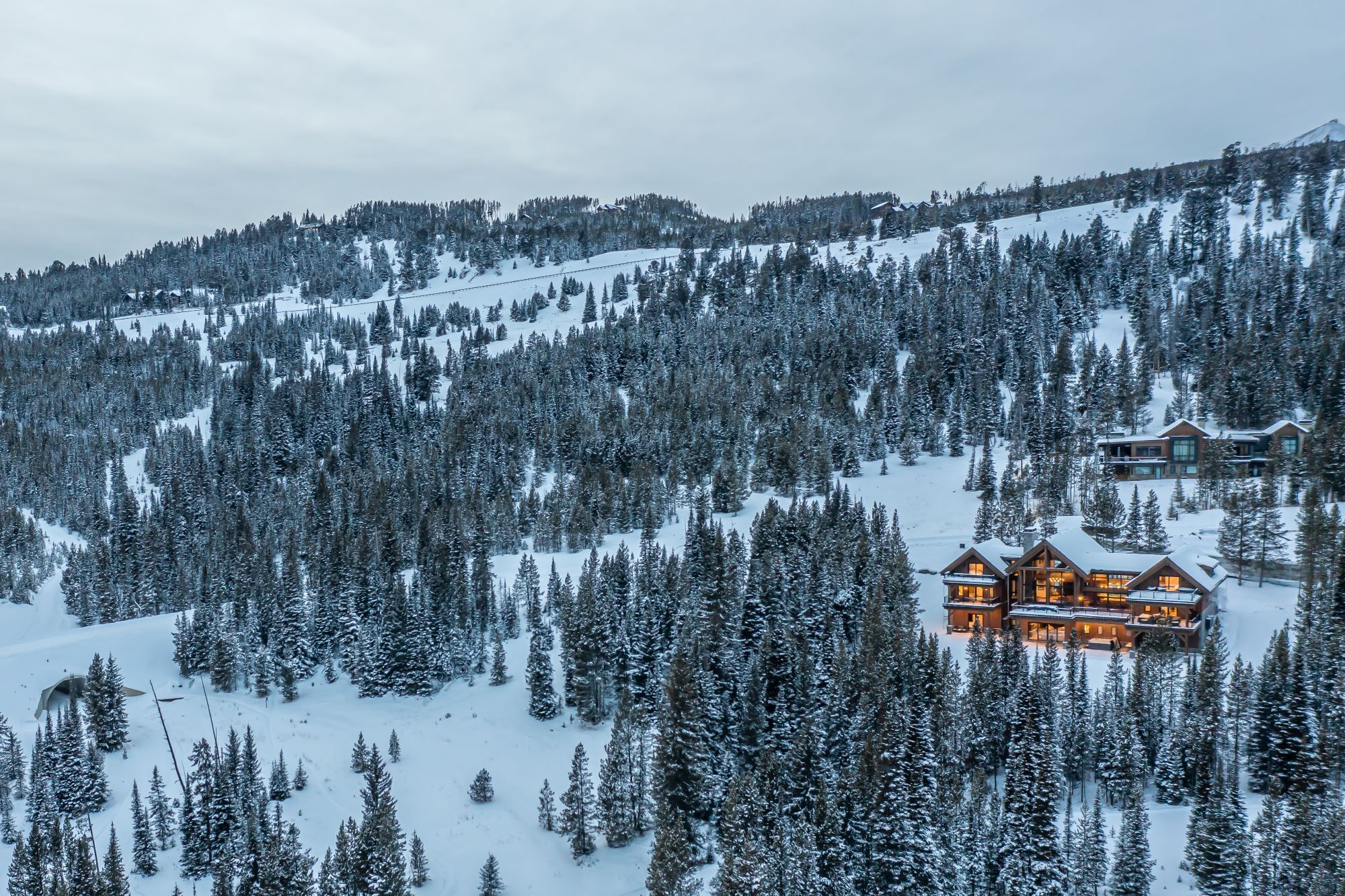 Un chalet enneigé doté de nombreuses pièces, entouré d’une forêt dense et adossé à une montagne couverte de neige.