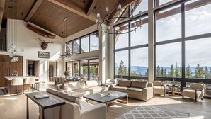 A spacious living area with a large window offering a mountain view, a dining area with a long table, and a kitchen with modern appliances. - Newly Built | Ski-in/Ski-out | Hot Tub (Big Sky)