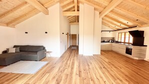 A spacious loft with high ceilings, exposed wooden beams, a grey sectional sofa, a small round table, and a kitchen area with a black range hood. - Ferienwohnung Mia, 100m², 2 Schlafzimmer, Wlan (Siegsdorf)