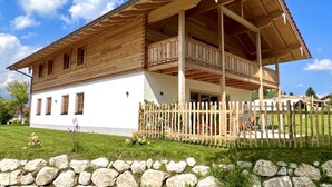Una casa de madera con balcón, un muro de piedra en primer plano y un patio cubierto de césped.